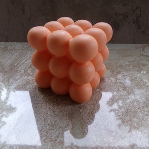 May include: A peach-coloured, cube-shaped stress ball made of soft, squishy material. The ball is made up of many small, round bumps.