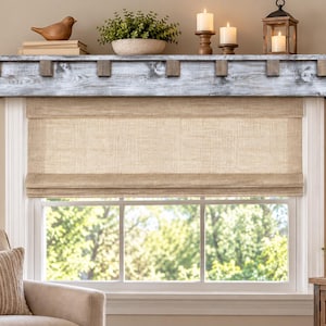 May include: A beige fabric roman shade installed over a window, featuring a textured weave. Above the window is a rustic, weathered gray wooden shelf with decorative block accents, supporting a bird figurine, books, a plant, and candles.