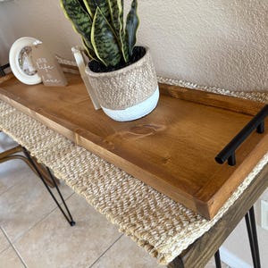 May include: A rectangular wooden tray with a medium brown finish and two matte black metal handles on the ends. The tray holds a white and tan textured planter containing a green snake plant and a small white circular frame with text.