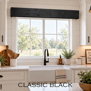 May include: A rectangular black wooden window valance with a visible wood grain texture mounted above a kitchen window. The valance features a dark, matte finish and spans the width of the window frame, positioned between two white kitchen cabinets.