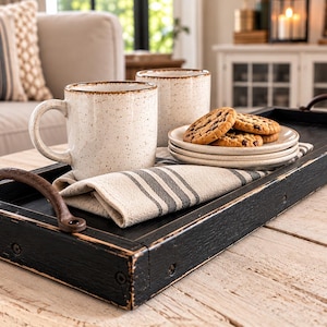 May include: A rectangular black wooden serving tray with distressed edges and two curved metal handles. Inside the tray are two speckled cream ceramic mugs, a stack of small plates with chocolate chip cookies, and a folded beige striped napkin.