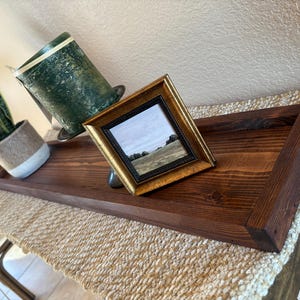 May include: A dark stained wooden tray with a gold framed picture and a green candle holder. A potted snake plant sits on the left side of the tray. The tray is on a woven runner.