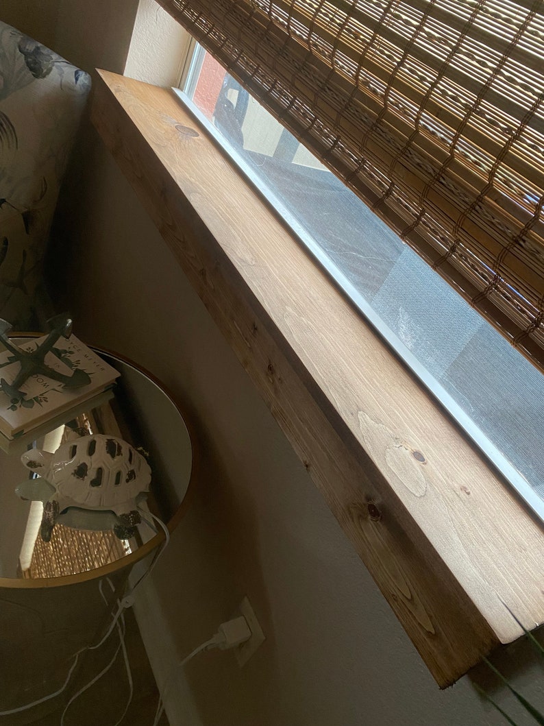 Puede incluir: Una estanter&iacute;a de madera marr&oacute;n claro est&aacute; montada sobre una ventana con una persiana de bamb&uacute;. La estanter&iacute;a est&aacute; inclinada hacia la c&aacute;mara y parece estar hecha de madera recuperada.