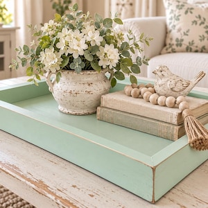 May include: A rectangular light green wooden tray containing a white ceramic pot with white flowers and green leaves, two stacked beige books, a small white bird figurine, and a string of wooden beads with a jute tassel attached to the books.