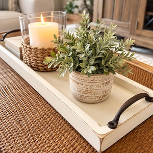 May include: A rectangular white wooden tray with dark metal handles holding a lit white pillar candle in a woven basket sleeve and a small green artificial plant in a distressed white ceramic pot with a textured, weathered finish.