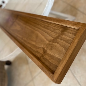 May include: A long, rectangular wooden shelf with a stained finish. The shelf has a raised edge around the perimeter.