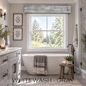 May include: A bathroom featuring a white freestanding bathtub, a gray vanity with black hardware, and a window with a whitewashed gray wooden valance. The room includes a small wooden stool, a textured towel, and framed botanical wall art.