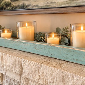May include: A long, rectangular, distressed teal wooden tray holding five clear glass cylindrical candle holders with lit white pillar candles, interspersed with artificial green eucalyptus leaves and small white berries.
