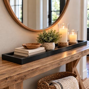 May include: A long, rectangular black tray holds two stacked beige books, a small wooden bowl, a potted green plant in a speckled ceramic pot, and two white pillar candles in clear glass cylinders with cork bases, arranged in a line.