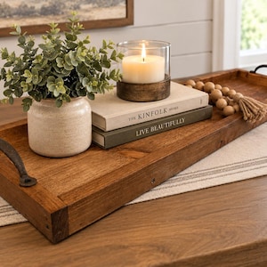 May include: A long rectangular brown wooden tray with dark metal handles. On the tray sits a small potted green plant, a white candle in a wooden base, two stacked books titled The Kinfolk Home and Live Beautifully, and wooden beads.
