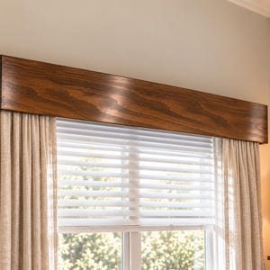 May include: A rectangular wooden window valance with a dark brown wood grain finish mounted above a window. The window features white horizontal blinds and is framed by floor-length, light beige curtains with a textured fabric appearance.