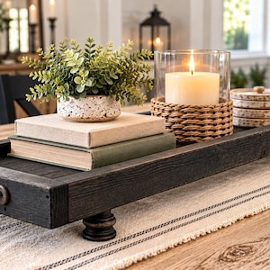 May include: A long, rectangular black wooden tray with dark metal handles and four small feet. The tray holds a potted green plant in a speckled white container, two stacked books, a lit candle in a glass holder with a woven base, and coasters.