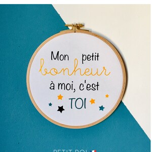 Pode incluir: Aro de bordado branco com o texto "Mon petit bonheur à moi, c'est TOI" em letras pretas e amarelas. O aro é rodeado por pequenas estrelas amarelas e azuis.