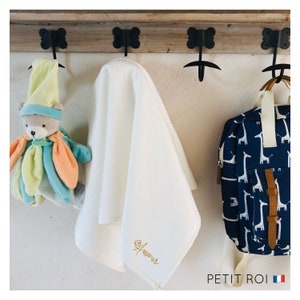 Puede incluir: Una manta de beb&eacute; blanca con la palabra "Amour" bordada en hilo dorado cuelga de un estante de madera con ganchos. Un animal de peluche y una mochila azul y blanca con estampado de jirafa cuelgan de los ganchos junto a la manta.