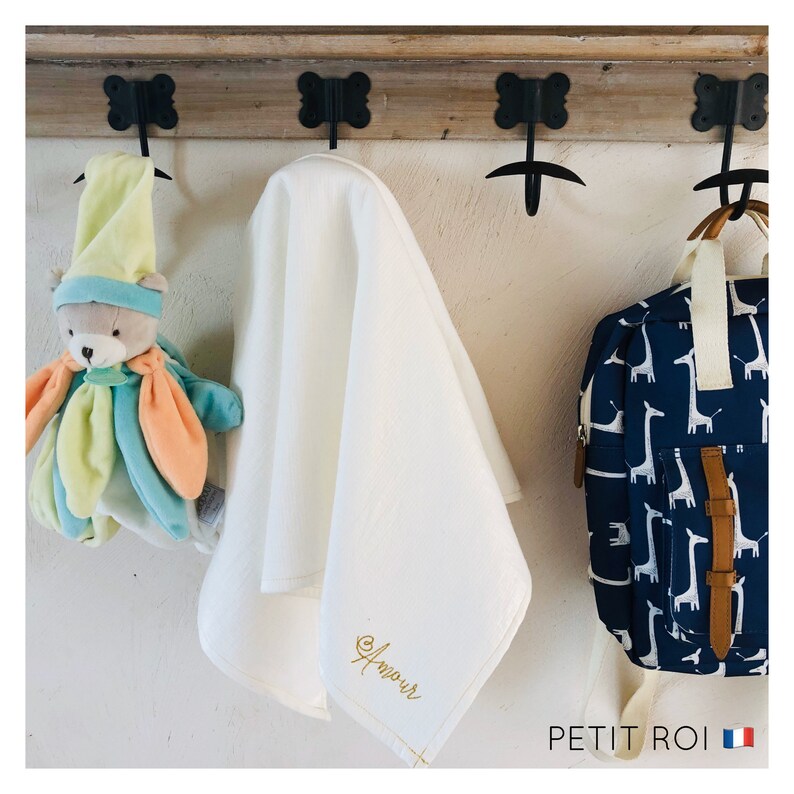 Puede incluir: Una manta de beb&eacute; blanca con la palabra "Amour" bordada en hilo dorado cuelga de un estante de madera con ganchos. Un animal de peluche y una mochila azul y blanca con estampado de jirafa cuelgan de los ganchos junto a la manta.