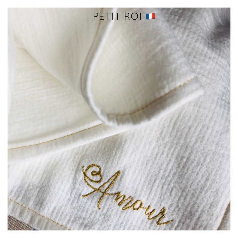 Puede incluir: Tela de algod&oacute;n blanca con la palabra "Amour" bordada en oro. La tela tiene una textura sutil y est&aacute; doblada sobre s&iacute; misma.