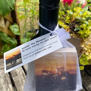 Op de afbeelding: Een donkere glazen wijnfles met een wit organza zakje met trekkoord om de hals gebonden. Het zakje bevat vierkante onderzetters met een afbeelding van wijnglazen bij zonsondergang. Een wit rechthoekig label aan het zakje vermeldt: Savour the Moment, 30 savvy sayings for wine lovers!, Compiled by Pink Gazelle Productions, Visit us at pinkgazelle.com.