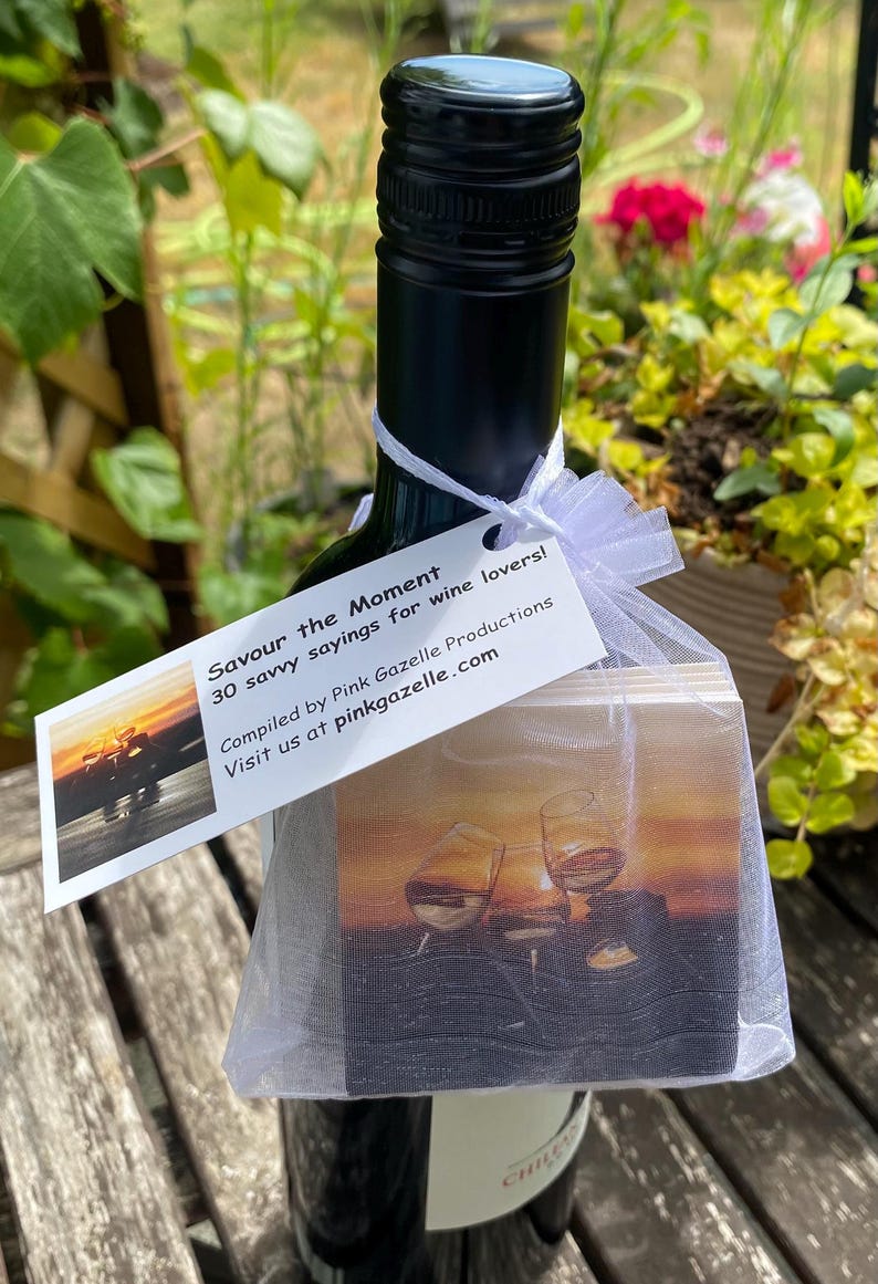 Op de afbeelding: Een donkere glazen wijnfles met een wit organza zakje met trekkoord om de hals gebonden. Het zakje bevat vierkante onderzetters met een afbeelding van wijnglazen bij zonsondergang. Een wit rechthoekig label aan het zakje vermeldt: Savour the Moment, 30 savvy sayings for wine lovers!, Compiled by Pink Gazelle Productions, Visit us at pinkgazelle.com.