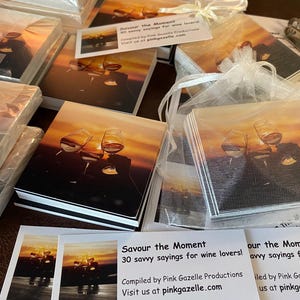 Op de afbeelding: Een stapel vierkante kaarten met een zonsondergangafbeelding van wijnglazen, verpakt in een wit organza zakje. Elke kaartenset bevat een label met de tekst Savour the Moment 30 savvy sayings for wine lovers! Compiled by Pink Gazelle Productions.