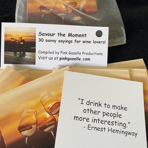 Op de afbeelding: Een set witte kaarten getiteld Savour the Moment 30 savvy sayings for wine lovers samengesteld door Pink Gazelle Productions. Elke kaart bevat een afbeelding van wijnglazen bij zonsondergang en een citaat, waaronder een van Ernest Hemingway.