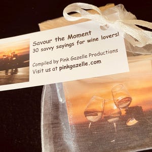 Op de afbeelding: Een wit organza zakje met trekkoord bevat kaarten met wijnafbeeldingen. Een wit label bevestigd met een lint toont de tekst: Savour the Moment, 30 savvy sayings for wine lovers! Compiled by Pink Gazelle Productions. Visit us at pinkgazelle.com.