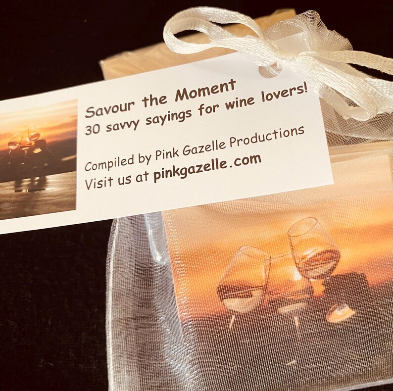 Op de afbeelding: Een wit organza zakje met trekkoord bevat kaarten met wijnafbeeldingen. Een wit label bevestigd met een lint toont de tekst: Savour the Moment, 30 savvy sayings for wine lovers! Compiled by Pink Gazelle Productions. Visit us at pinkgazelle.com.