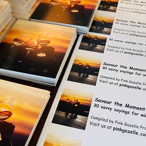 Op de afbeelding: Stapels vierkante boekjes met een zonsondergangfoto van twee klinkende wijnglazen. De tekst luidt: Savour the Moment, 30 savvy sayings for wine lovers! Compiled by Pink Gazelle Productions. Visit us at pinkgazelle.com.