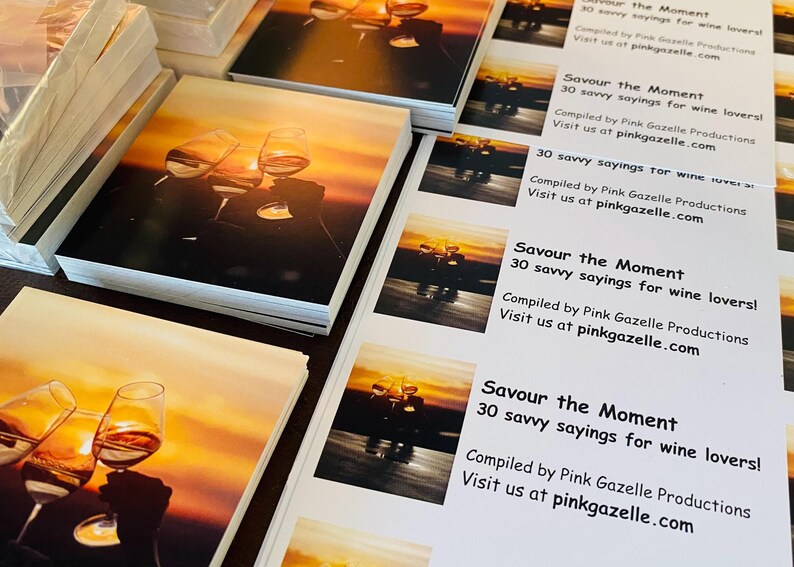 Op de afbeelding: Stapels vierkante boekjes met een zonsondergangfoto van twee klinkende wijnglazen. De tekst luidt: Savour the Moment, 30 savvy sayings for wine lovers! Compiled by Pink Gazelle Productions. Visit us at pinkgazelle.com.