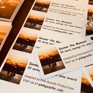 Op de afbeelding: Een stapel rechthoekige witte kaarten met een zonsondergangfoto van wijnglazen en de tekst: Savour the Moment, 30 savvy sayings for wine lovers!, Compiled by Pink Gazelle Productions, Visit us at pinkgazelle.com. Een metalen perforator is zichtbaar.
