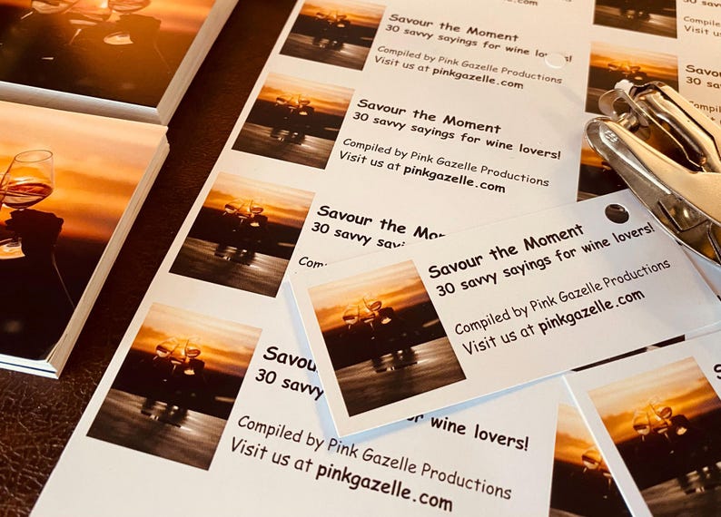 Op de afbeelding: Een stapel rechthoekige witte kaarten met een zonsondergangfoto van wijnglazen en de tekst: Savour the Moment, 30 savvy sayings for wine lovers!, Compiled by Pink Gazelle Productions, Visit us at pinkgazelle.com. Een metalen perforator is zichtbaar.