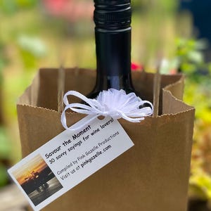 Op de afbeelding: Een bruine papieren cadeauzak met een donkere glazen fles met een zwarte schroefdop. Een wit rechthoekig label is met een wit tule lint aan de zak bevestigd. Het label bevat de tekst "Savour the Moment 30 savvy sayings for wine lovers!"