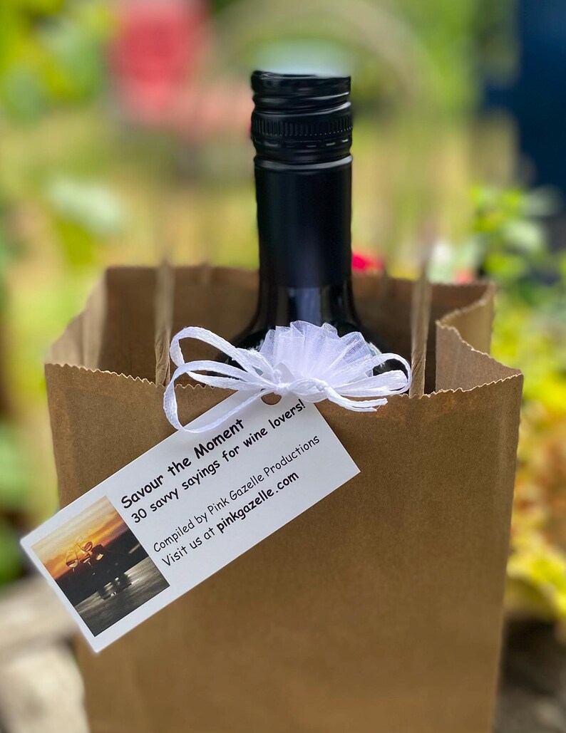 Op de afbeelding: Een bruine papieren cadeauzak met een donkere glazen fles met een zwarte schroefdop. Een wit rechthoekig label is met een wit tule lint aan de zak bevestigd. Het label bevat de tekst "Savour the Moment 30 savvy sayings for wine lovers!"