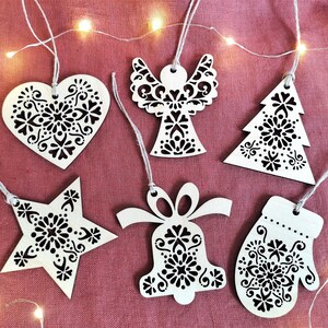 Puede incluir: Seis adornos navideños de madera blanca con intrincados diseños florales negros. Los adornos tienen forma de corazón, ángel, estrella, campana, árbol de Navidad y manopla.