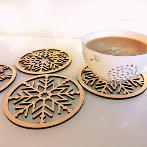 Puede incluir: Cuatro posavasos de madera con un diseño de copo de nieve. Los posavasos están hechos de madera marrón claro y tienen un diseño de copo de nieve recortado. Una taza blanca con un líquido marrón está sentada en uno de los posavasos.