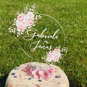 May include: Clear acrylic cake topper with a floral design and the text "Gabriele in Jonas".