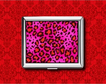 May include: A silver cigarette case with a vibrant pink leopard print design. The case features a repeating pattern of orange and black spots against a bright pink background. The case is set against a red patterned background.