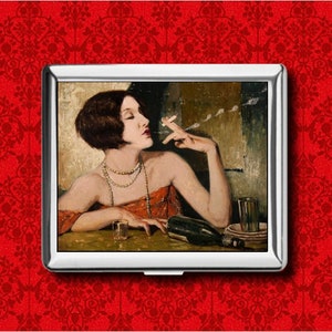 May include: A silver cigarette case featuring a vintage painting of a woman smoking. The artwork depicts a woman with short dark hair, wearing a red dress and multiple necklaces, seated at a table with a drink and a bottle.