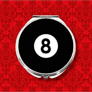 May include: A round, silver compact mirror with a black face featuring a white number 8, set against a red background with a floral pattern. The compact is closed and centered.