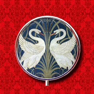 May include: A round, silver-coloured compact with a blue and green design featuring two white swans facing each other. The swans are surrounded by stylised reeds and leaves.