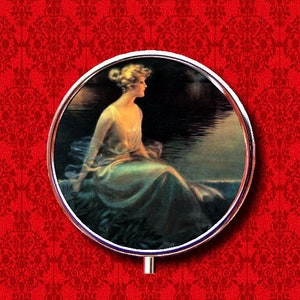May include: A vintage illustration of a woman in a flowing blue dress sitting by a lake at dusk. The woman has blonde hair and is looking out at the water. The image is framed in a silver circle.