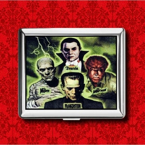 May include: A silver metal cigarette case featuring a classic horror movie poster design. The image includes Dracula, Frankenstein, The Mummy, and The Wolfman, with a green background and lightning bolts. The case is set against a red patterned background.