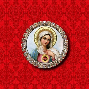 May include: A round, rhinestone-encrusted brooch featuring a depiction of the Virgin Mary with a heart in her hands. The image is set against a white background with a gold halo around the Virgin Mary's head.
