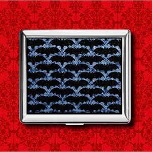 May include: A silver metal cigarette case with a repeating pattern of black bat silhouettes against a blue and white background. The case is rectangular and sits against a red floral patterned background.