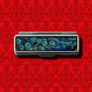 Puede incluir: Una caja de pastillas de metal plateado con un diseño de plumas de pavo real. Las plumas son azules, verdes y doradas.