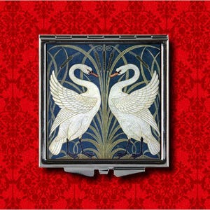 May include: A silver compact mirror with a blue and green design featuring two white swans facing each other. The swans are surrounded by stylized reeds and leaves.