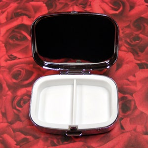 May include: A silver pill box with a mirrored lid and two compartments. The pill box is open and the compartments are empty.
