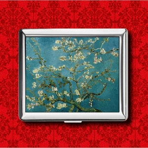 May include: A silver cigarette case displaying a print of Vincent van Gogh's "Almond Blossoms". The image shows a blossoming almond tree against a blue sky. The case is rectangular and set against a red patterned background.