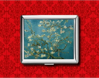 May include: A silver cigarette case featuring a print of Vincent van Gogh's "Almond Blossoms" painting. The artwork depicts a blossoming almond tree against a blue sky. The case is rectangular and set against a red patterned background.