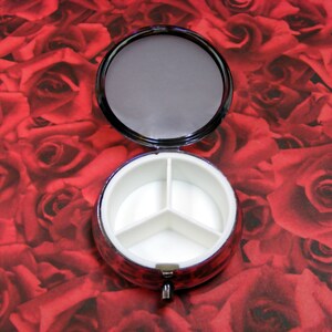 May include: A round, silver pill box with a red rose pattern. The box has three compartments inside.