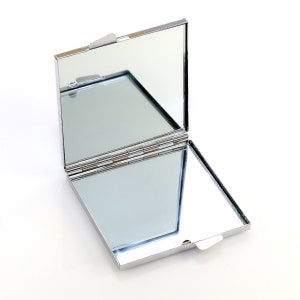 May include: A silver compact mirror with a hinged lid. The mirror is open, revealing a reflective surface.